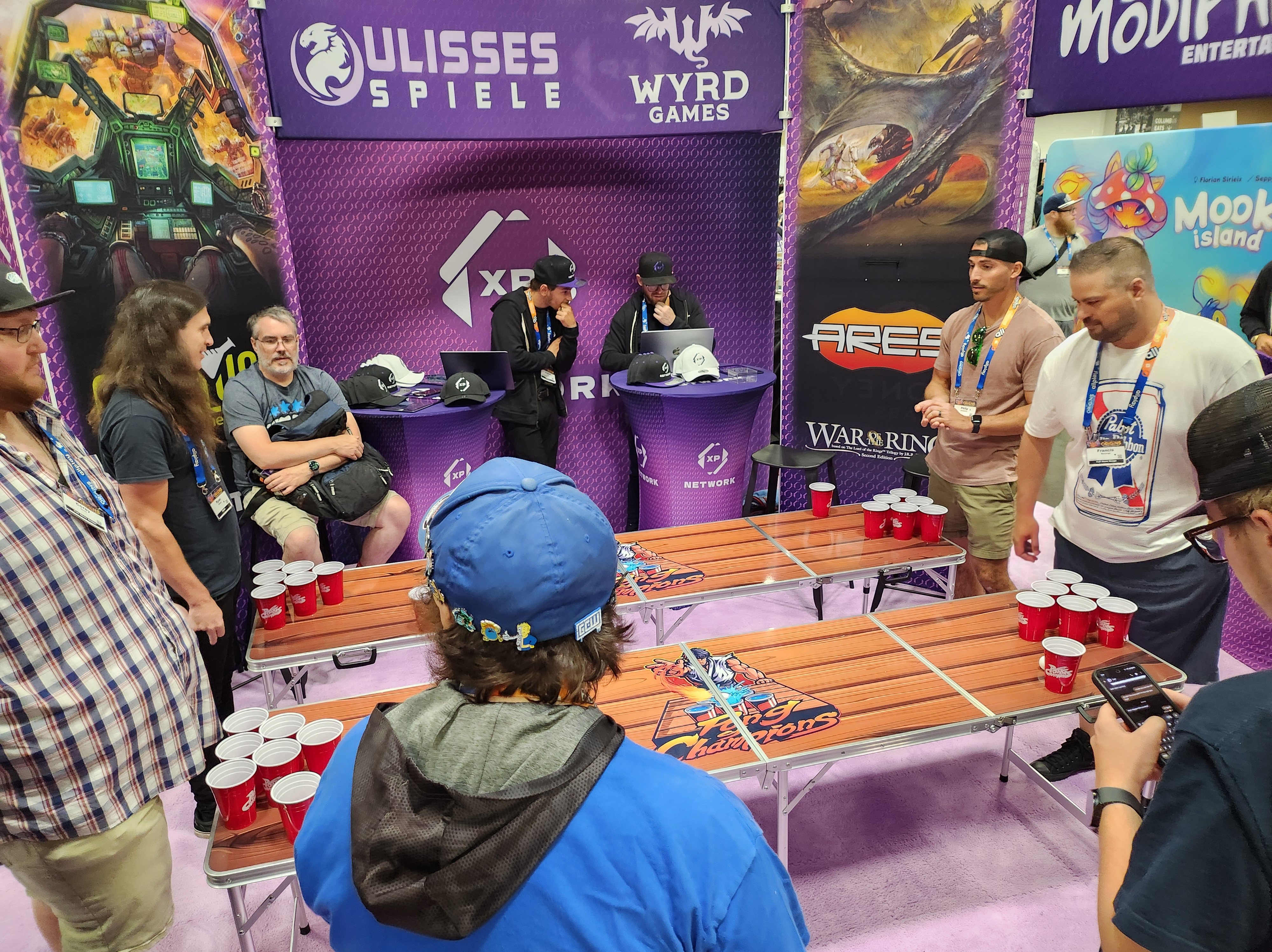 Inside the XP Network booth, two groups are facing off in a 1v1 game of 6-cup pong. Each group has players competing in a head-to-head match, creating a lively and competitive atmosphere.
